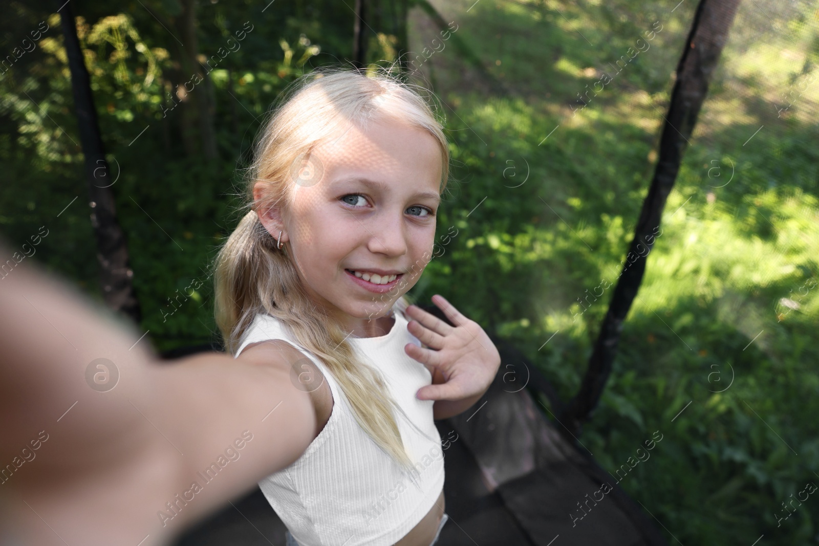 Photo of Happy little girl taking selfie and waving hello on black trampoline outdoors. Space for text