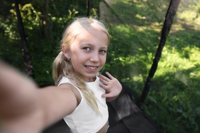 Photo of Happy little girl taking selfie and waving hello on black trampoline outdoors. Space for text