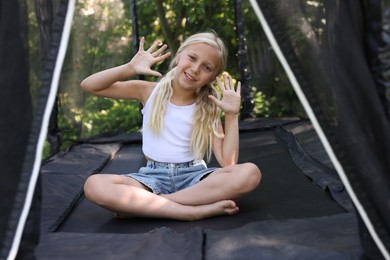 Photo of Happy little girl waving hello and sitting on black trampoline outdoors