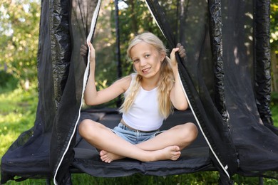 Photo of Happy little girl sitting on black trampoline outdoors