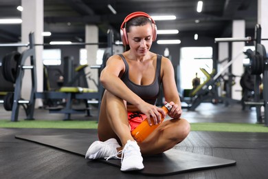 Photo of Athletic woman in headphones with bottle resting on yoga mat in gym
