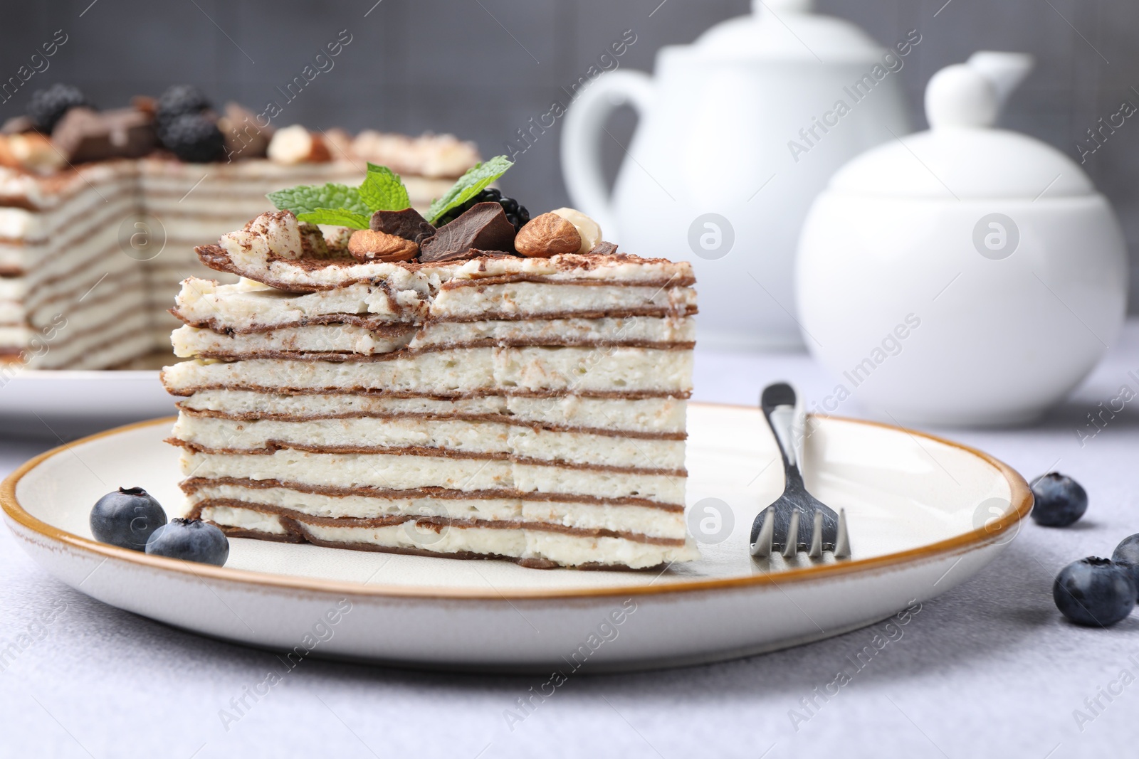 Photo of Piece of tasty crepe cake with cocoa powder, chocolate, nuts and blackberry served on light table, closeup