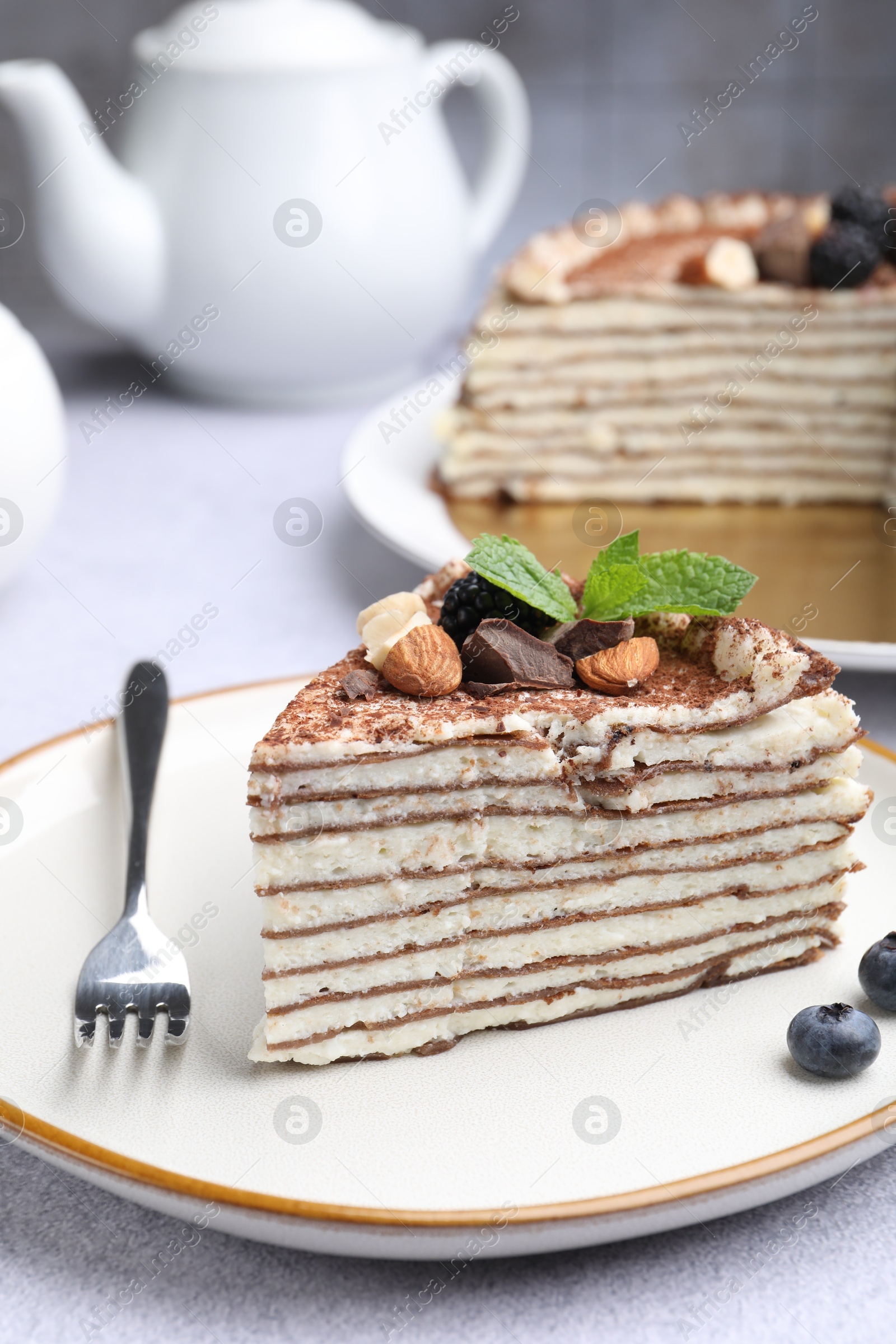 Photo of Piece of tasty crepe cake with cocoa powder, chocolate, nuts and blackberry served on light table, closeup