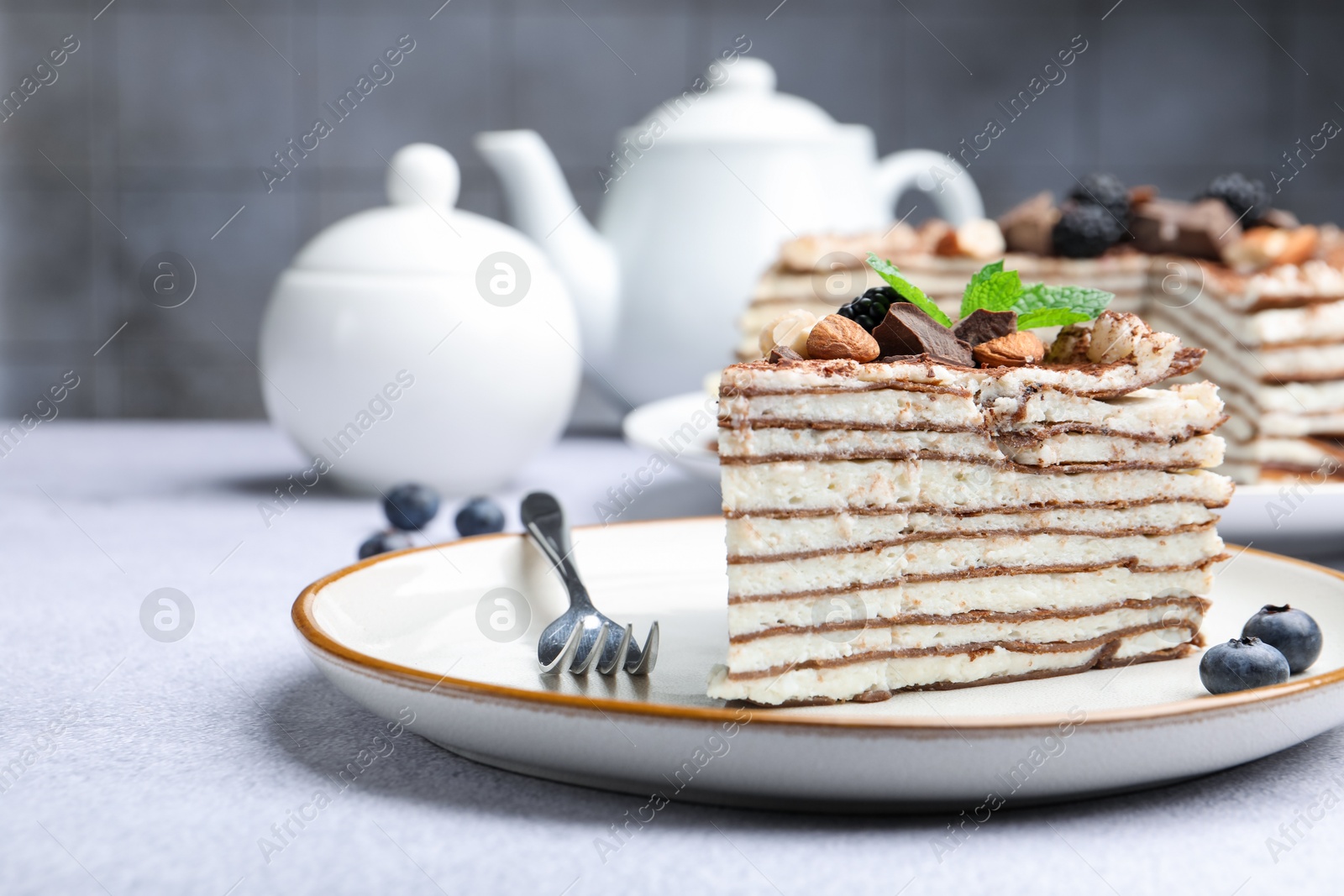 Photo of Piece of tasty crepe cake with cocoa powder, chocolate, nuts and blackberry served on light table, closeup