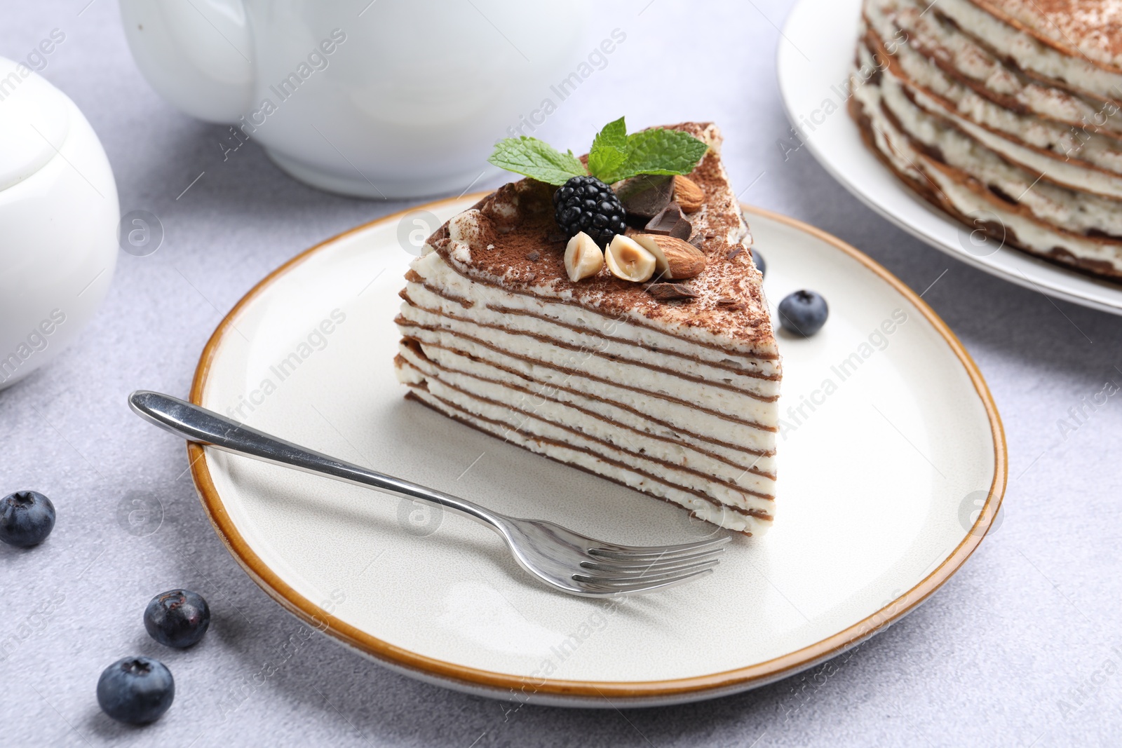 Photo of Piece of tasty crepe cake with cocoa powder, chocolate, nuts and blackberry served on light table, closeup