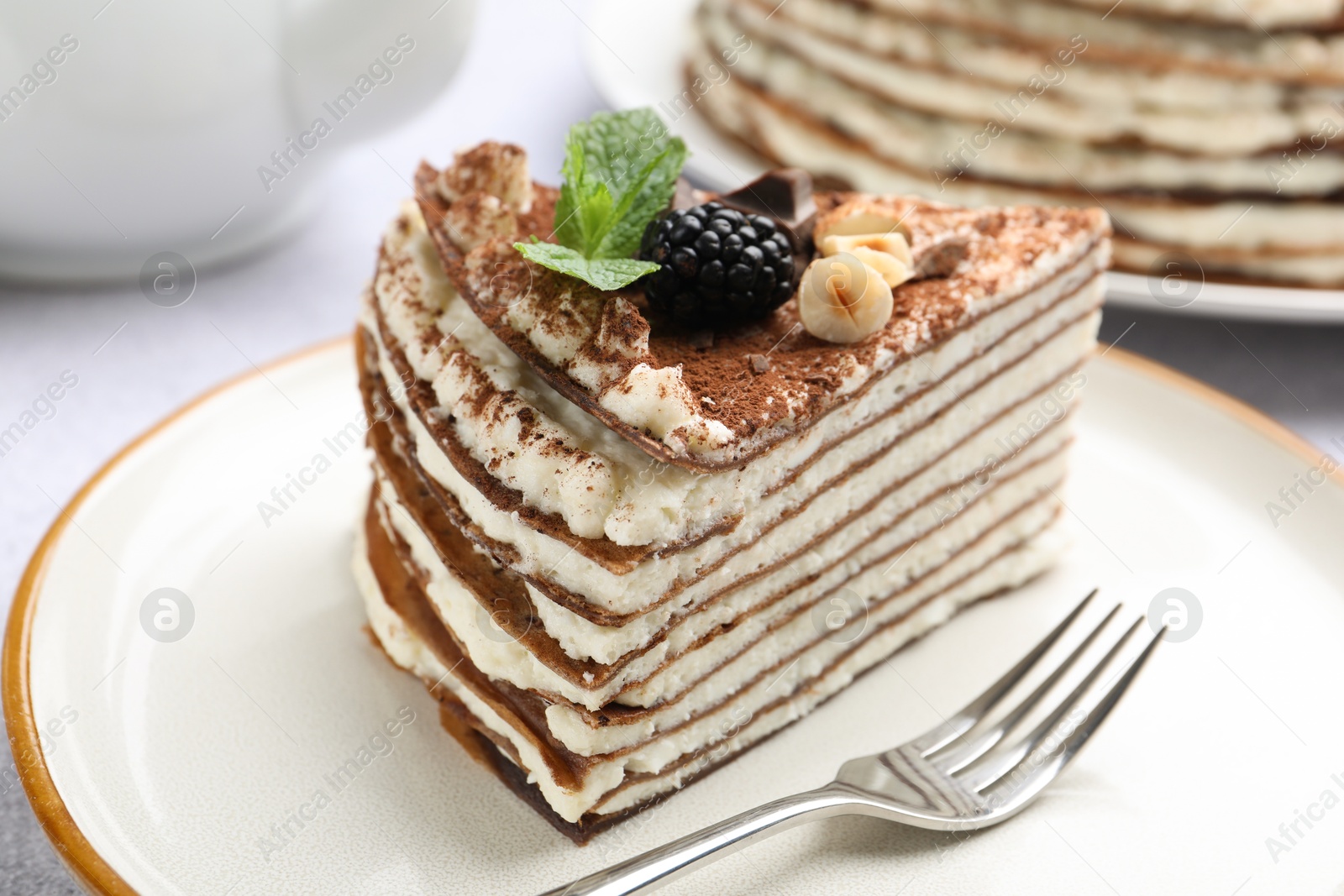 Photo of Piece of tasty crepe cake with cocoa powder, chocolate, nuts and blackberry served on light table, closeup