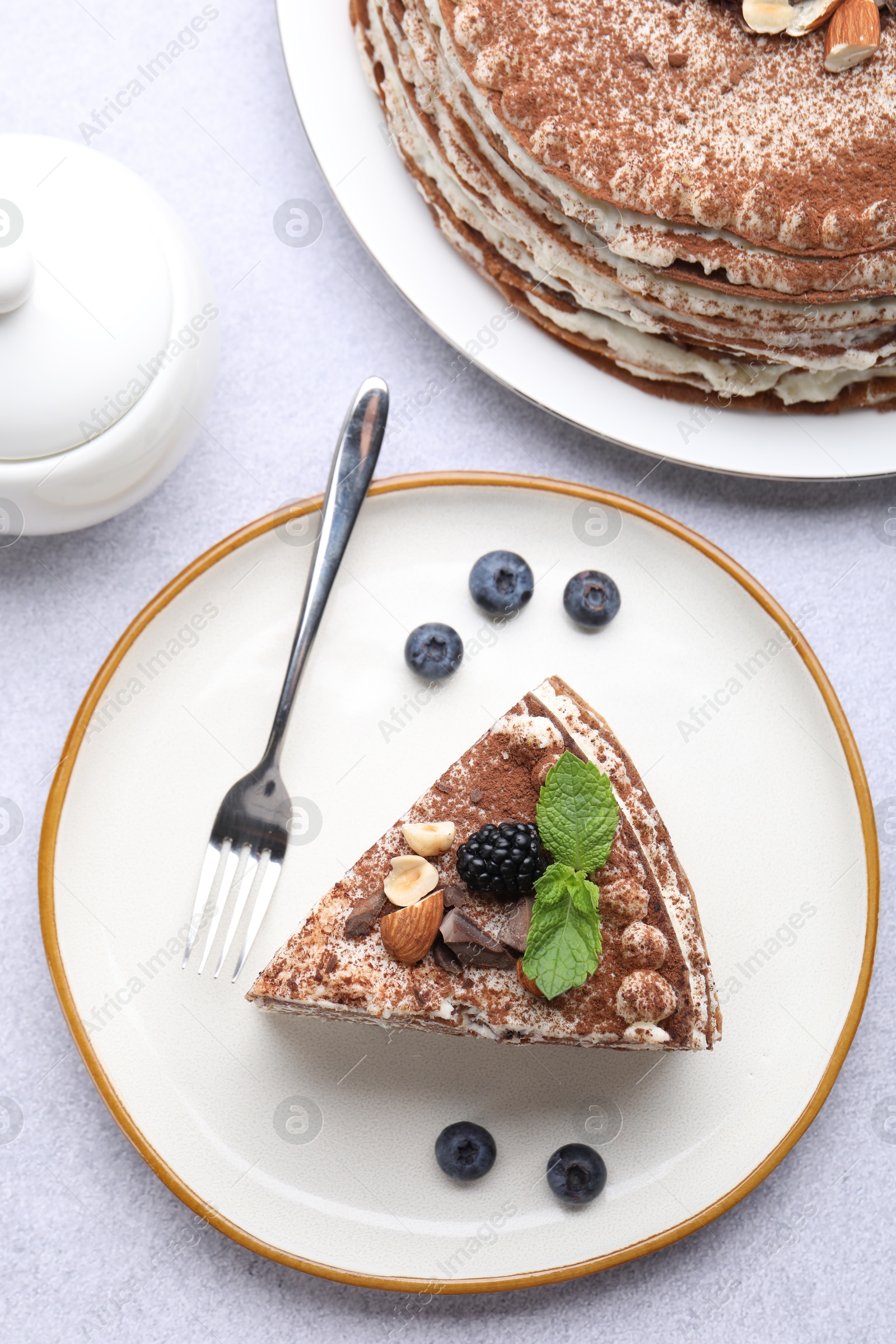 Photo of Piece of tasty crepe cake with cocoa powder, chocolate, nuts and blackberry served on light table, flat lay