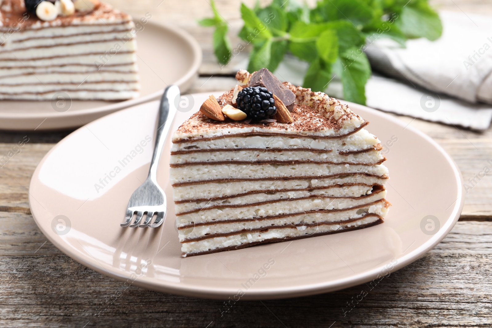 Photo of Piece of tasty crepe cake with cocoa powder, chocolate, nuts and blackberry served on wooden table, closeup