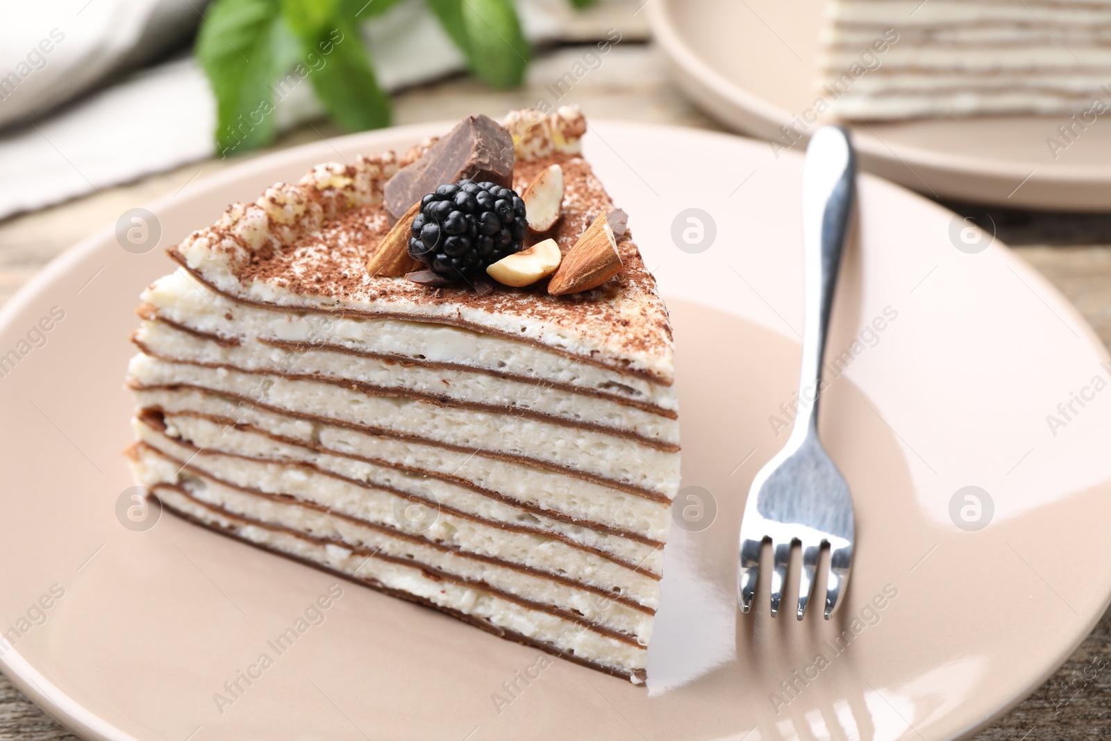 Photo of Piece of tasty crepe cake with cocoa powder, chocolate, nuts and blackberry served on wooden table, closeup