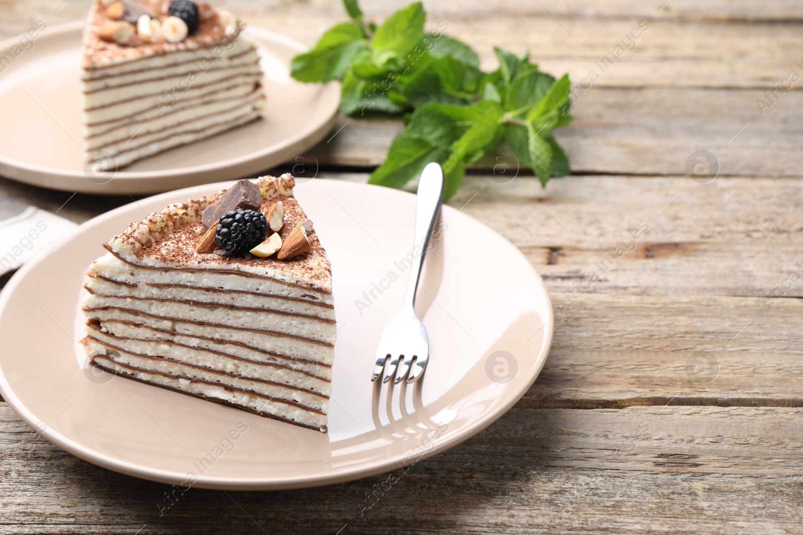 Photo of Piece of tasty crepe cake with cocoa powder, chocolate, nuts and blackberry served on wooden table, closeup