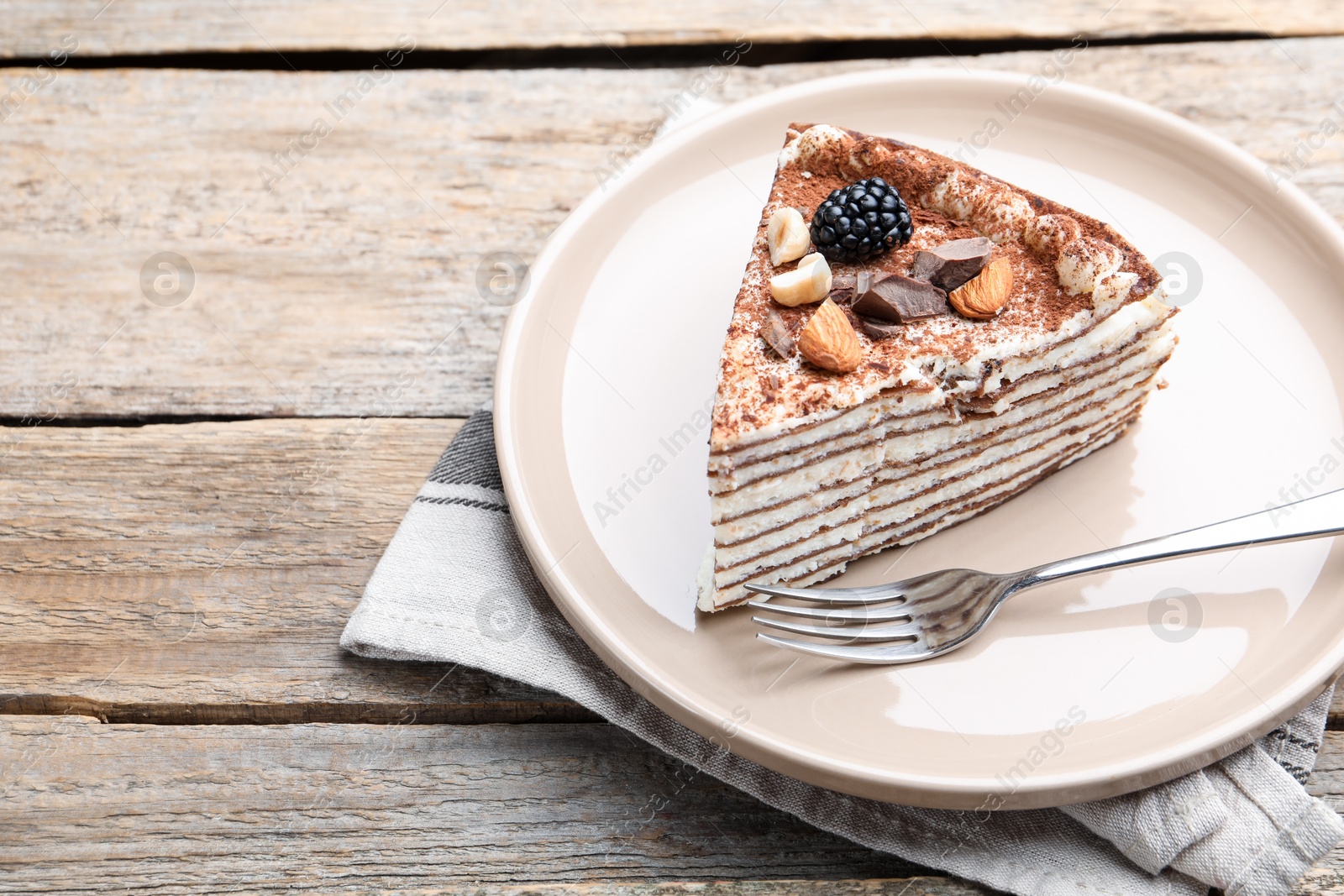 Photo of Piece of tasty crepe cake with cocoa powder, chocolate, nuts and blackberry served on wooden table, closeup. Space for text