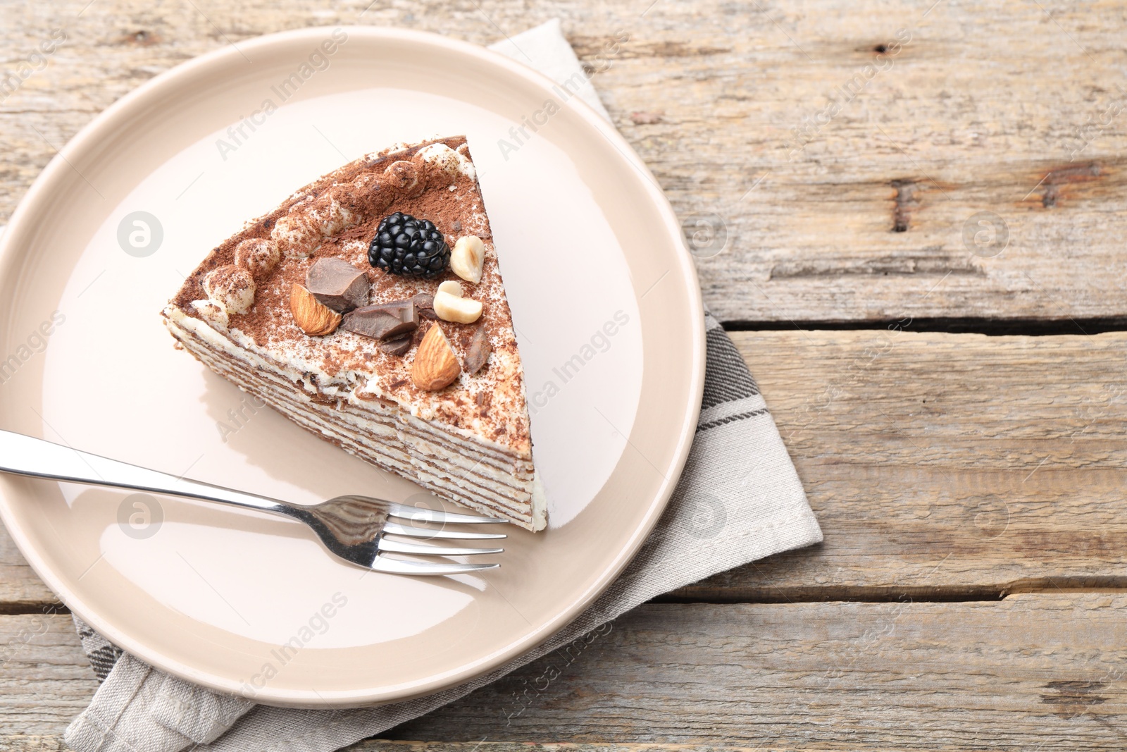 Photo of Piece of tasty crepe cake with cocoa powder, chocolate, nuts and blackberry served on wooden table, closeup. Space for text