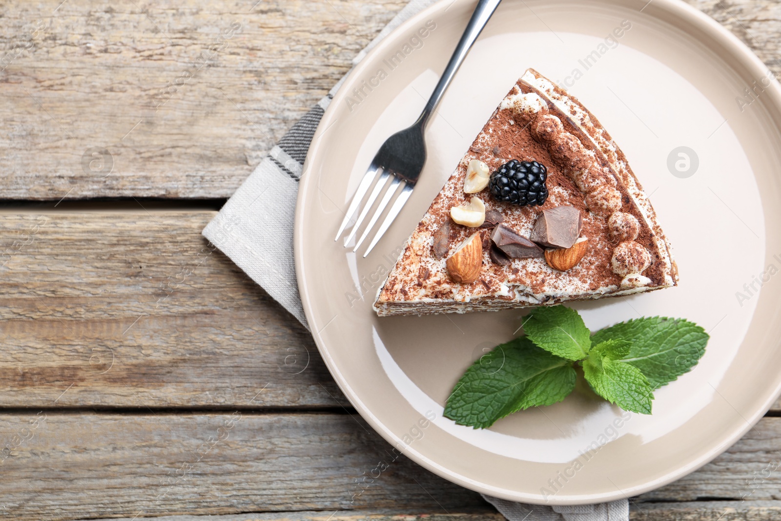 Photo of Piece of tasty crepe cake with cocoa powder, chocolate, nuts and blackberry served on wooden table, top view. Space for text
