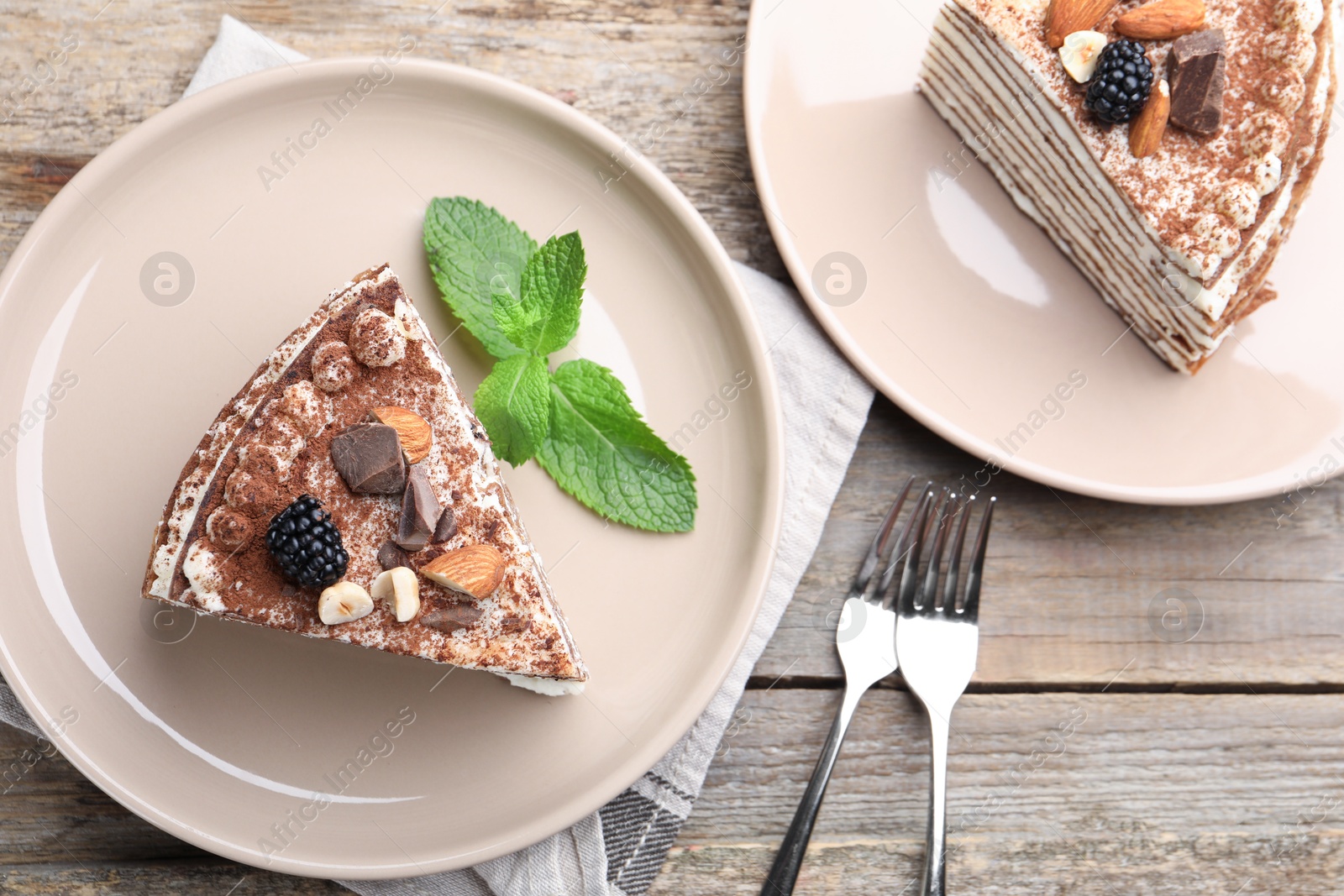 Photo of Piece of tasty crepe cake with cocoa powder, chocolate, nuts and blackberries served on wooden table, flat lay