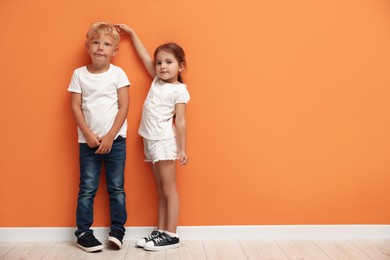 Photo of Cute little girl and boy measuring their heights near orange wall. Space for text