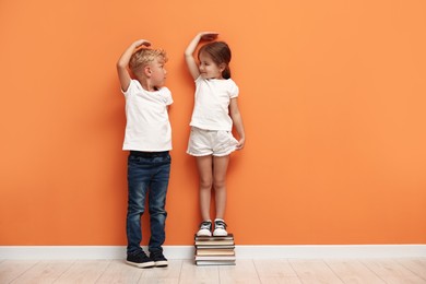 Photo of Cute little girl and boy measuring their heights near orange wall. Space for text