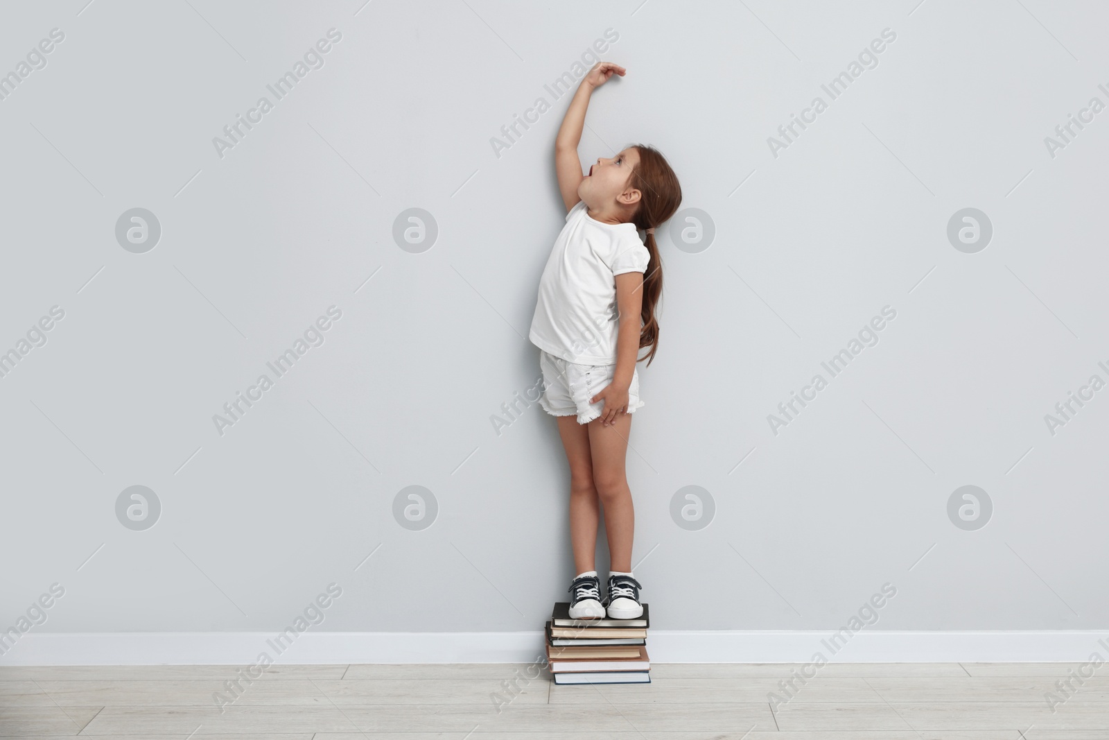 Photo of Cute little girl on stack of books measuring her height near light wall