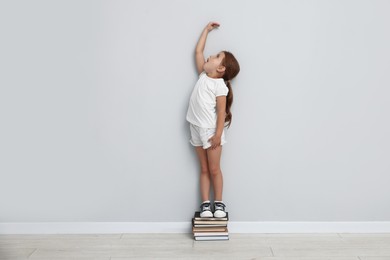 Photo of Cute little girl on stack of books measuring her height near light wall