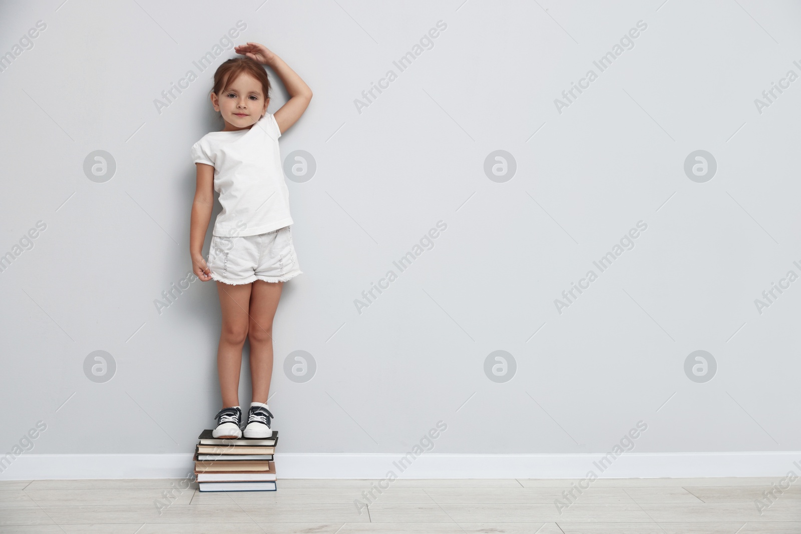 Photo of Cute little girl on stack of books measuring her height near light wall. Space for text