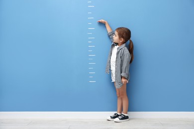 Photo of Cute little girl measuring her height near blue wall. Space for text
