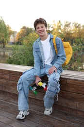 Photo of Happy teenage boy with backpack and penny board near lake outdoors