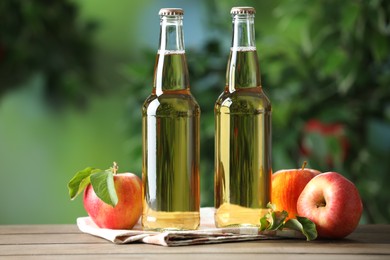 Photo of Delicious cider in glass bottles and apples on wooden table outdoors