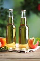 Photo of Delicious cider in glass bottles and apples on wooden table outdoors