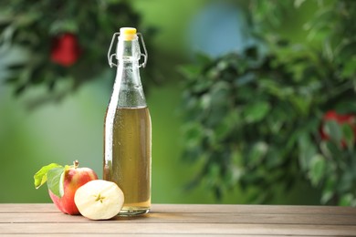 Photo of Delicious cider in glass bottle and apples on wooden table outdoors, space for text