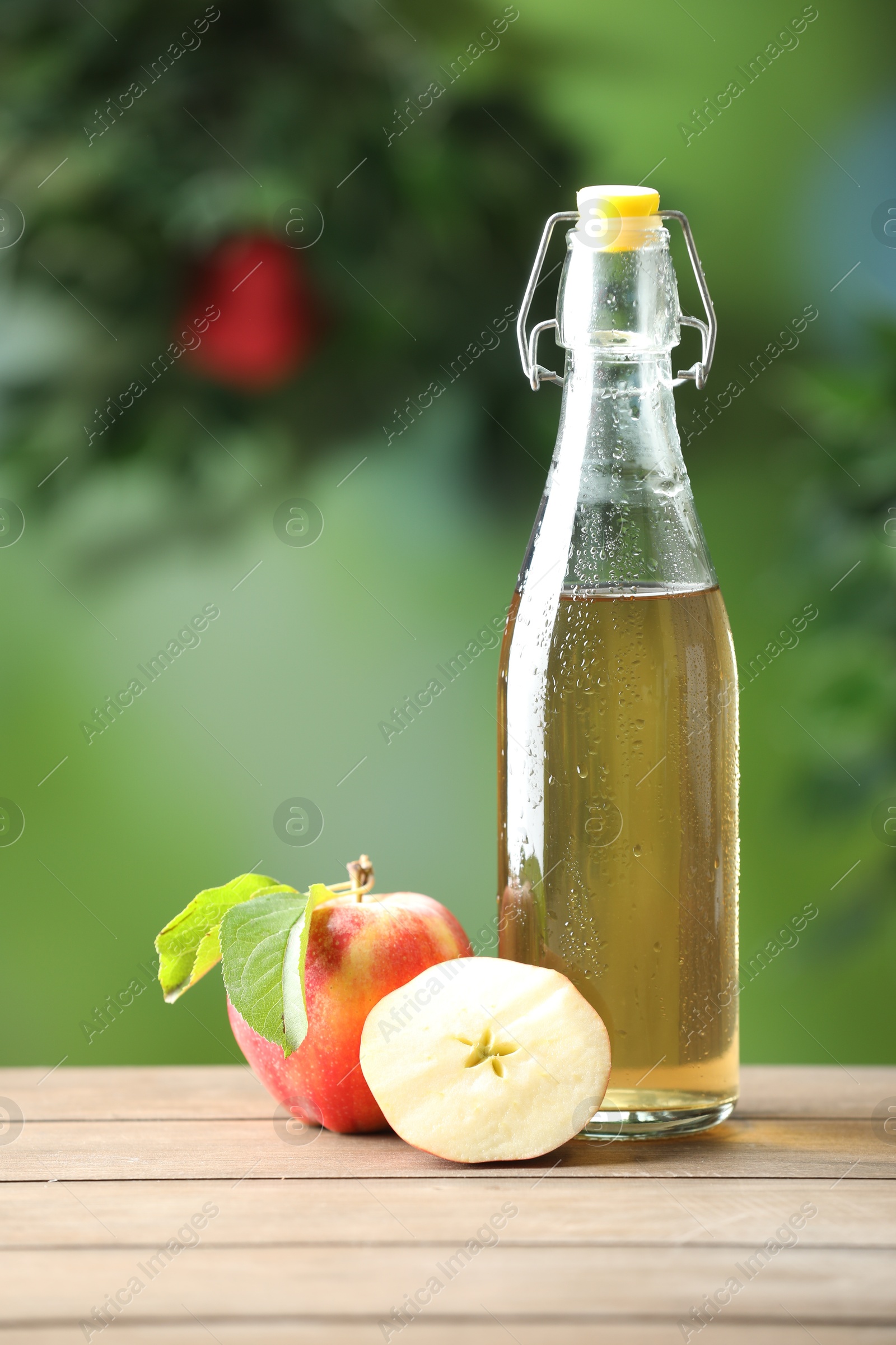 Photo of Delicious cider in glass bottle and apples on wooden table outdoors