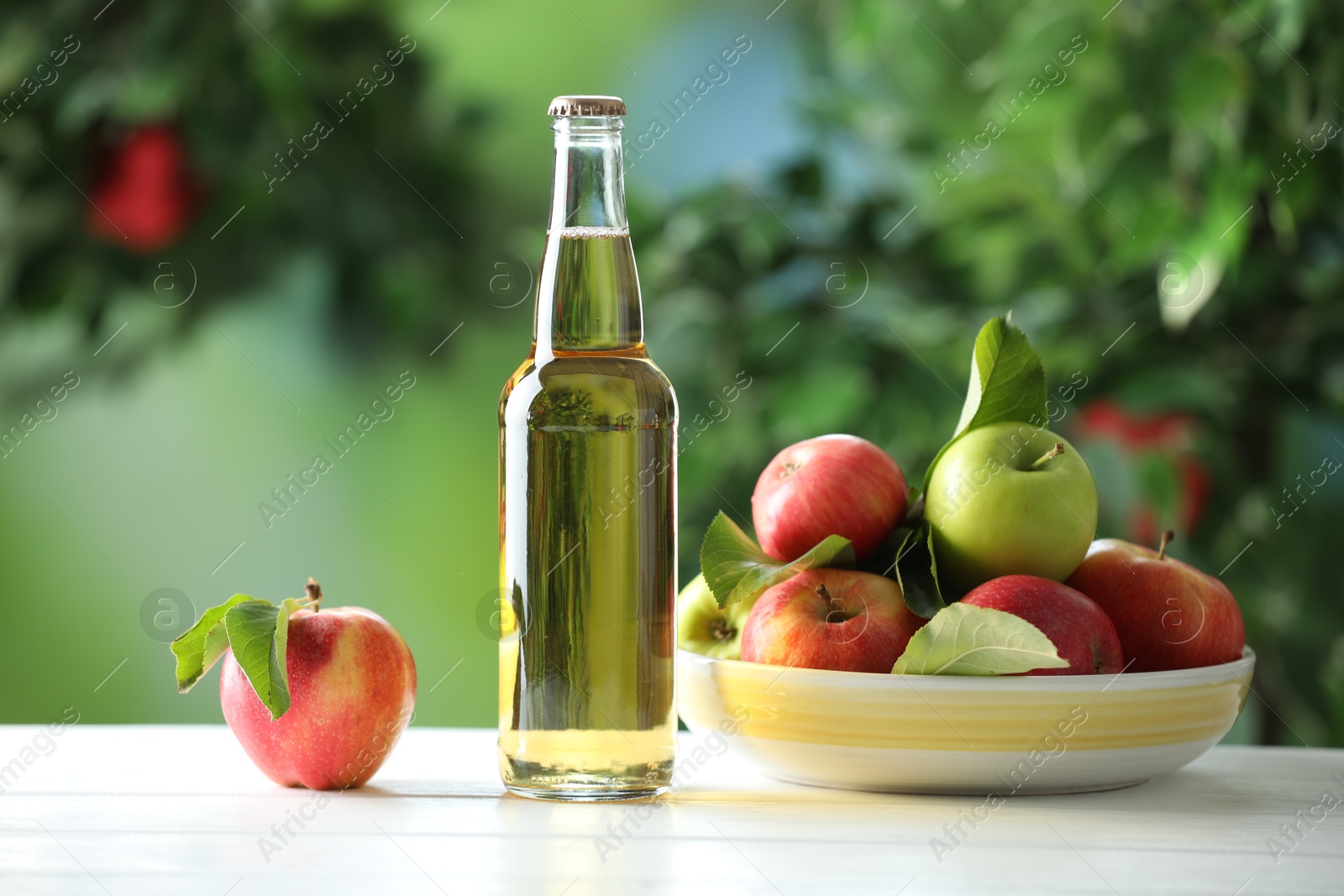Photo of Delicious cider in glass bottle and apples on white wooden table outdoors