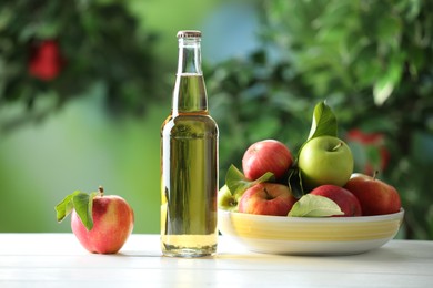 Photo of Delicious cider in glass bottle and apples on white wooden table outdoors
