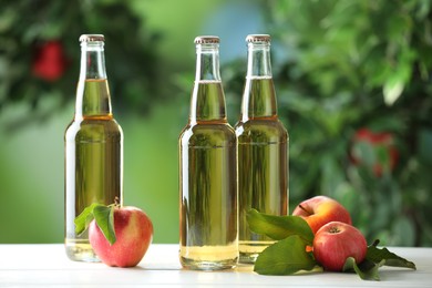 Photo of Delicious cider in glass bottles and apples on white wooden table outdoors