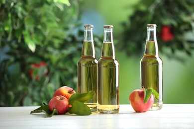 Photo of Delicious cider in glass bottles and apples on white wooden table outdoors