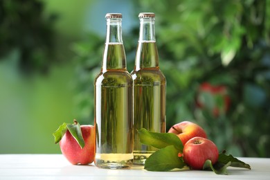 Photo of Delicious cider in glass bottles and apples on white wooden table outdoors