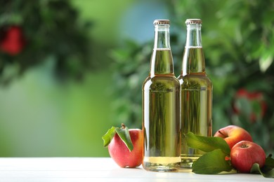 Photo of Delicious cider in glass bottles and apples on white wooden table outdoors, space for text