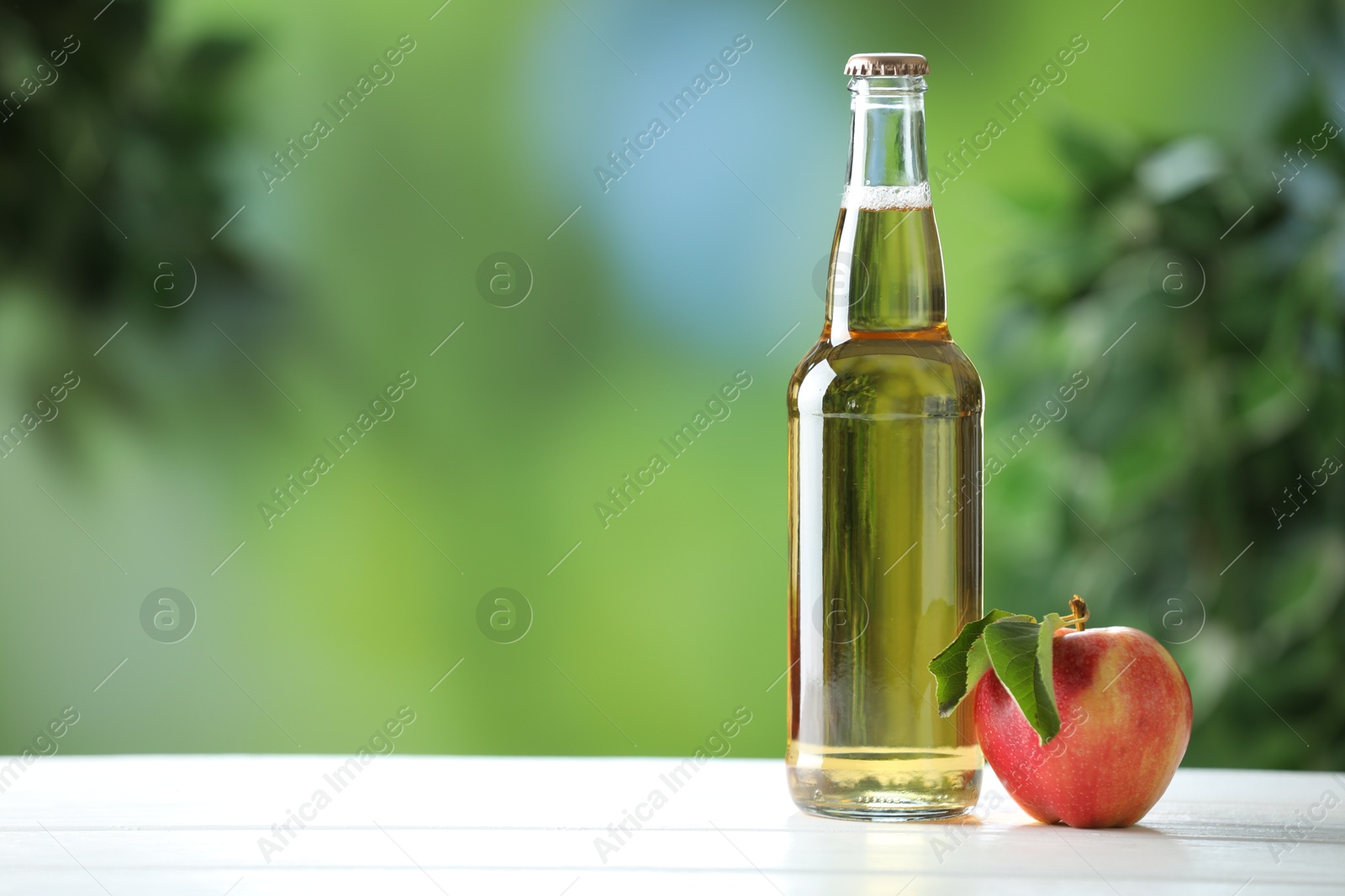 Photo of Delicious cider in glass bottle and apple on white wooden table outdoors, space for text