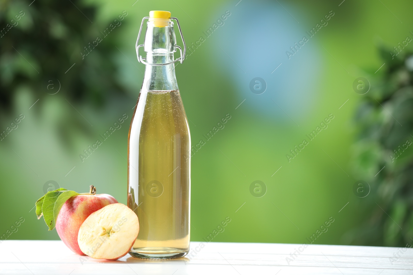 Photo of Delicious cider in glass bottle and apples on white wooden table outdoors, space for text