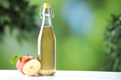 Photo of Delicious cider in glass bottle and apples on white wooden table outdoors, space for text