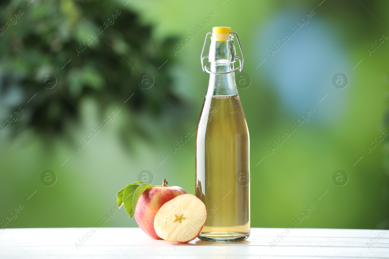 Photo of Delicious cider in glass bottle and apples on white wooden table outdoors
