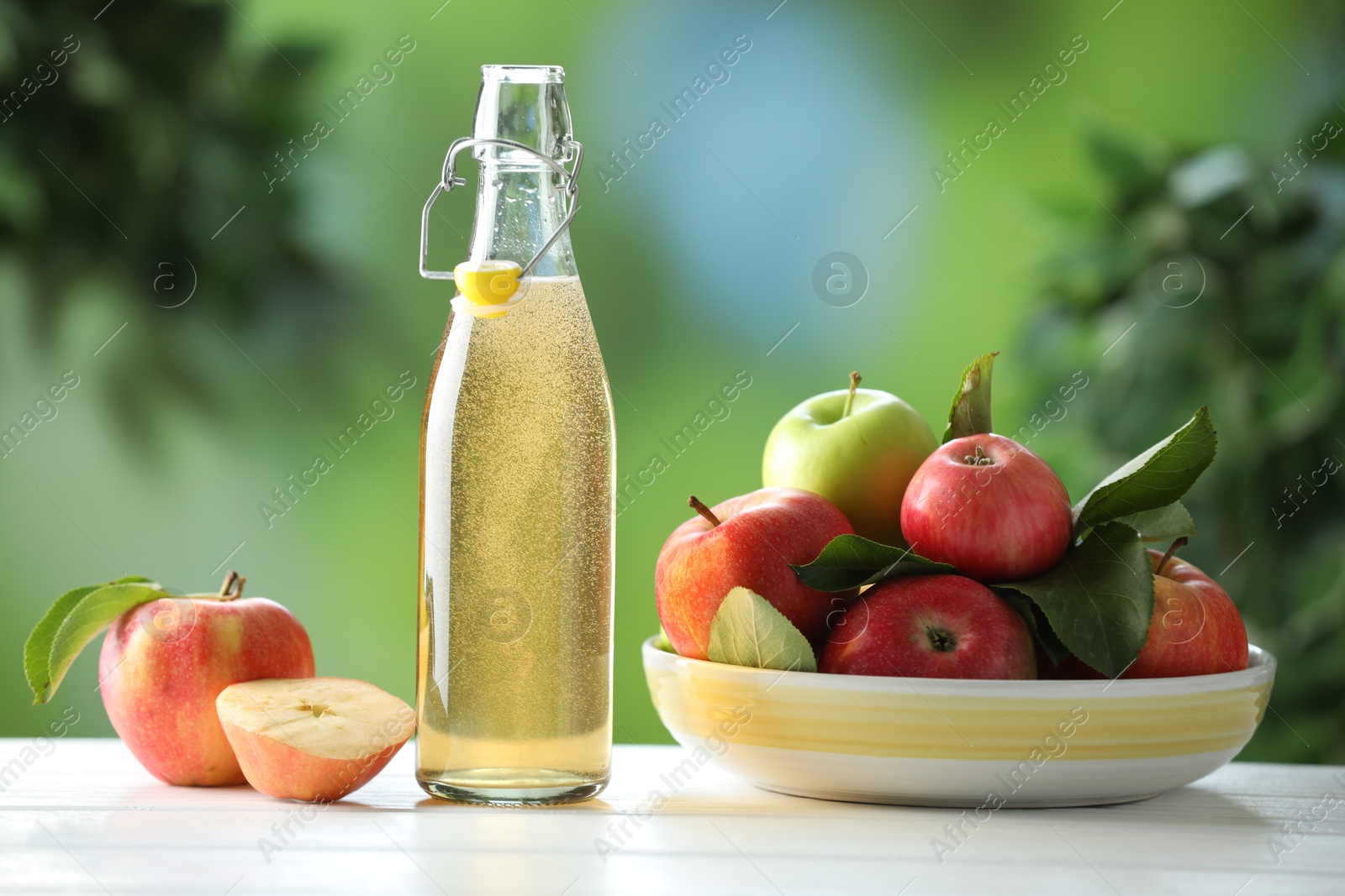 Photo of Delicious cider in glass bottle and apples on white wooden table outdoors