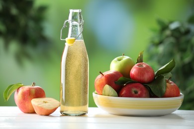 Photo of Delicious cider in glass bottle and apples on white wooden table outdoors