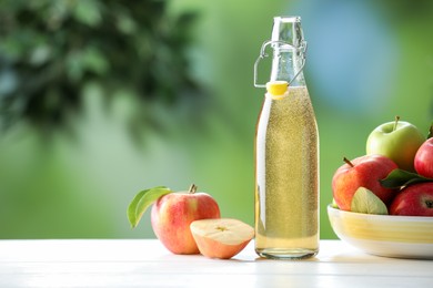 Photo of Delicious cider in glass bottle and apples on white wooden table outdoors, space for text