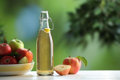 Photo of Delicious cider in glass bottle and apples on white wooden table outdoors, space for text