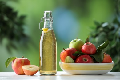 Photo of Delicious cider in glass bottle and apples on white wooden table outdoors