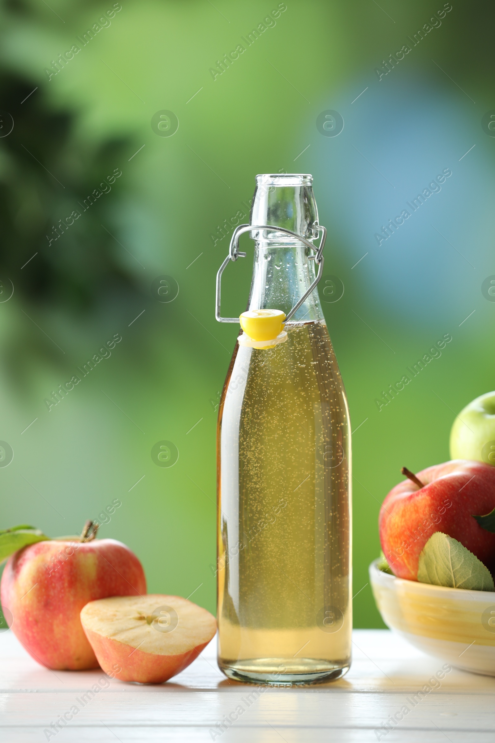 Photo of Delicious cider in glass bottle and apples on white wooden table outdoors