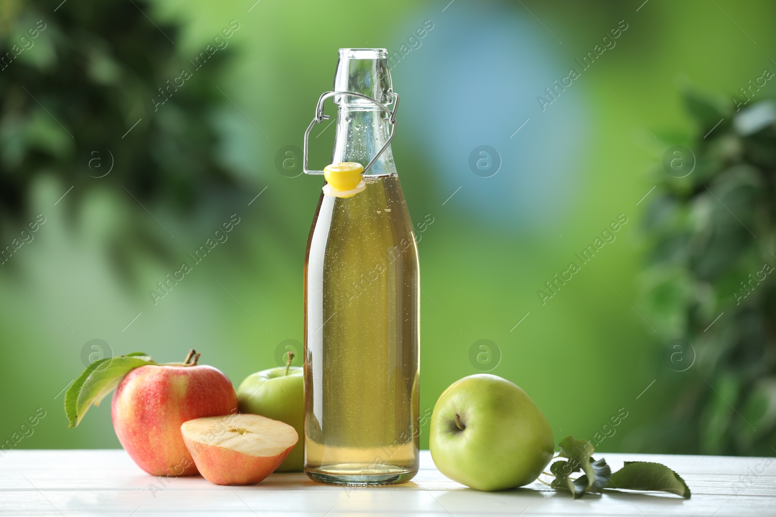 Photo of Delicious cider in glass bottle and apples on white wooden table outdoors