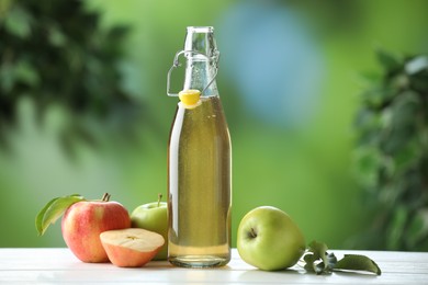 Photo of Delicious cider in glass bottle and apples on white wooden table outdoors