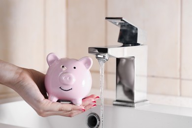 Photo of Water saving concept. Woman with piggy bank and water flowing out of tap indoors, closeup