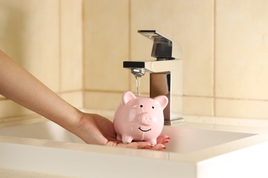 Photo of Water saving concept. Woman holding piggy bank under tap water indoors, closeup