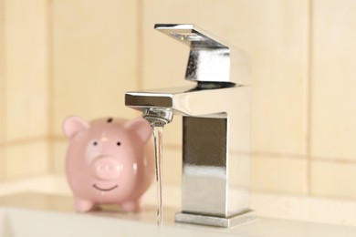 Photo of Water saving concept. Stream of water flowing from tap and piggy bank indoors, selective focus